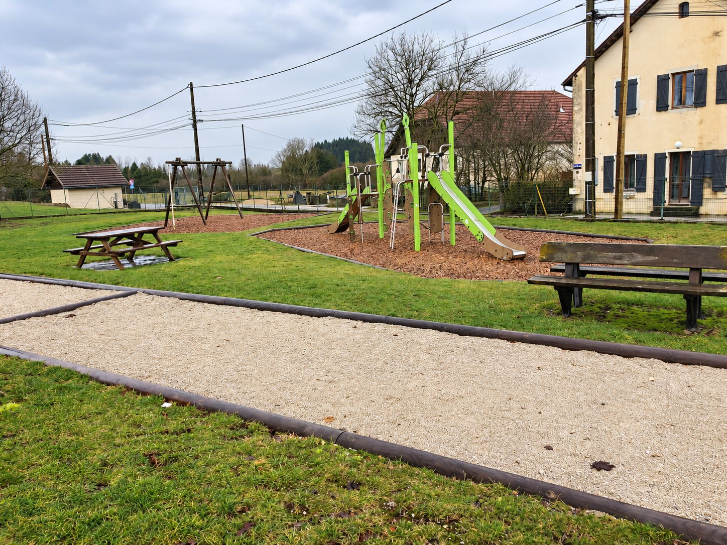Une aire de jeux avec toboggan vert, table de pique-nique et bancs, à côté d'un terrain de pétanque gravillonné, devant des maisons de village.