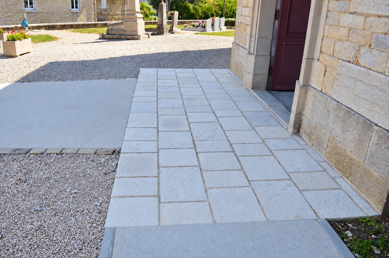 Une allée en dalles de pierre claire borde un mur en pierre et mène à une porte bordeaux, près d'un cimetière et d'un sol gravillonné.
