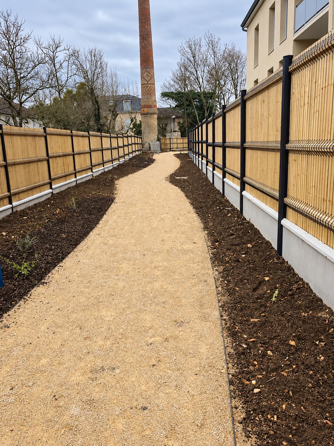 Une allée de gravier clair serpente entre deux clôtures en bois sur muret de béton, bordée de terre fraîchement remuée, avec une ancienne cheminée en briques au fond.
