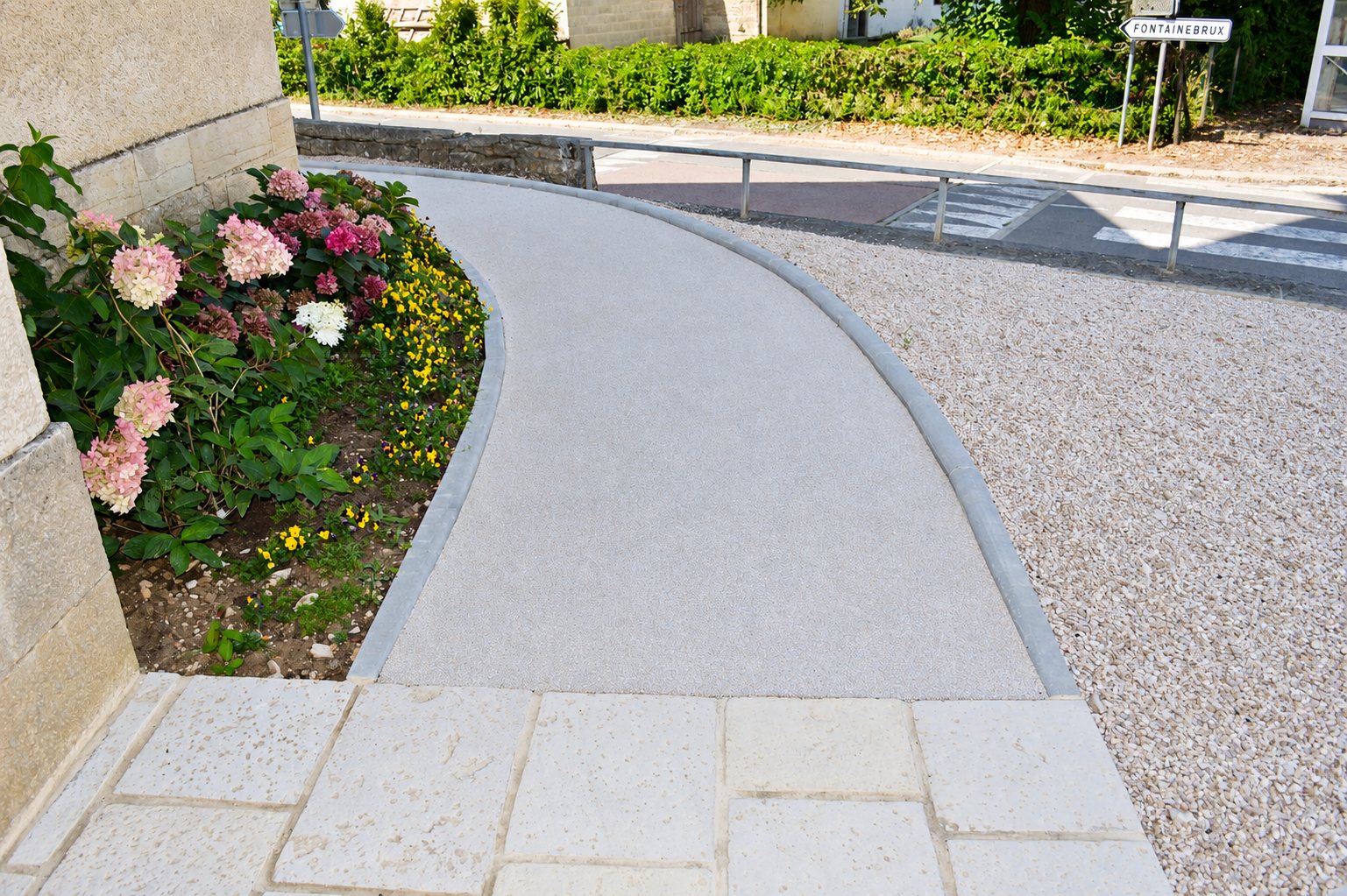 Une allée piétonne courbe en béton clair bordée d'un massif d'hortensias, avec des dalles en pierre au premier plan et du gravier à droite.