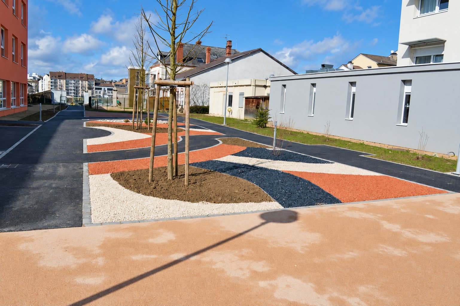 Espace extérieur aménagé avec des bandes de gravillons orange, blancs et noirs, de jeunes arbres tuteurés et des allées en enrobé entre plusieurs bâtiments.