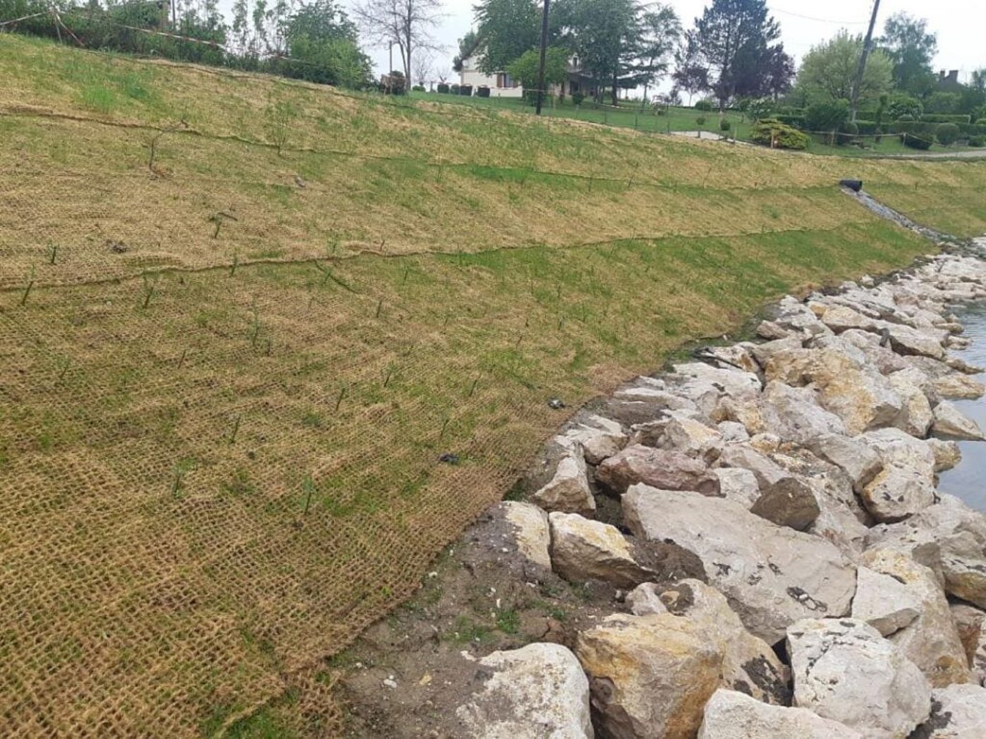 Talus de berge recouvert d'un filet de fibres naturelles où l'herbe commence à pousser, avec un enrochement de gros blocs de pierre au bord de l'eau.