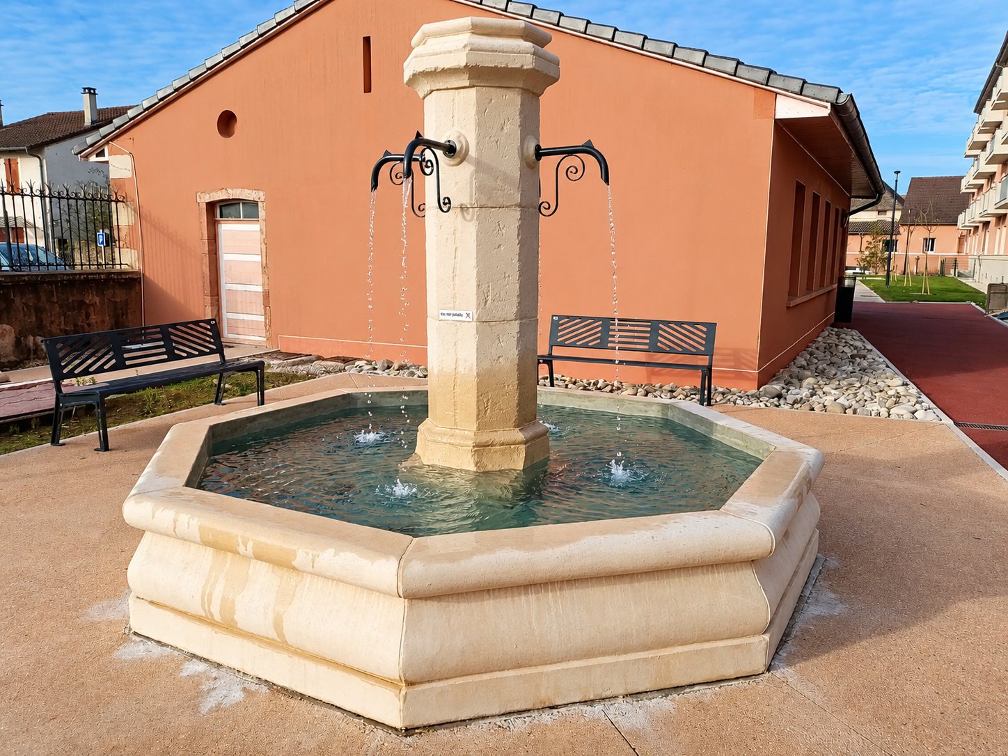 Une fontaine en pierre claire à bassin octogonal et colonne centrale, avec deux bancs noirs devant un bâtiment couleur saumon.