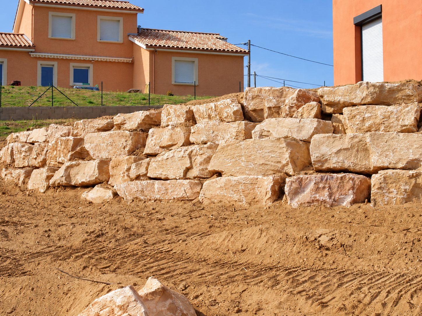 Un mur de soutènement en gros blocs de pierre beige empilés sur un terrain de terre sableuse, avec des maisons en crépi orangé à l'arrière-plan.