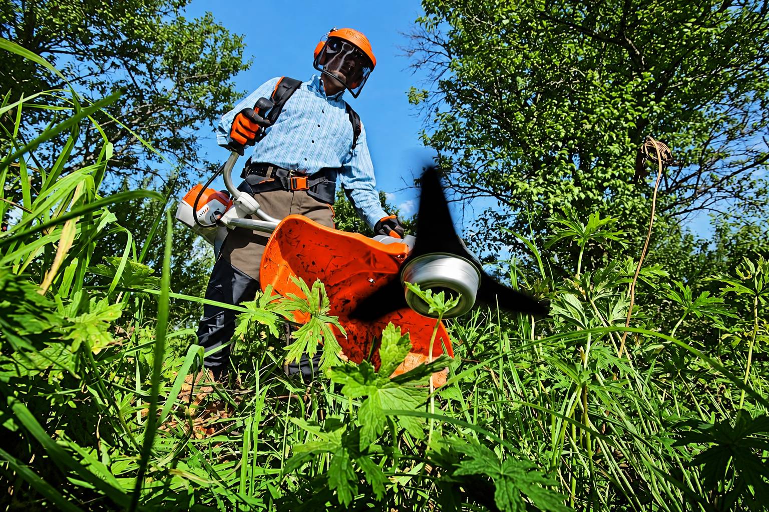 Vu depuis le sol, un homme casqué, avec visière et gants orange, utilise une débroussailleuse au milieu de hautes herbes et de feuillages verts.