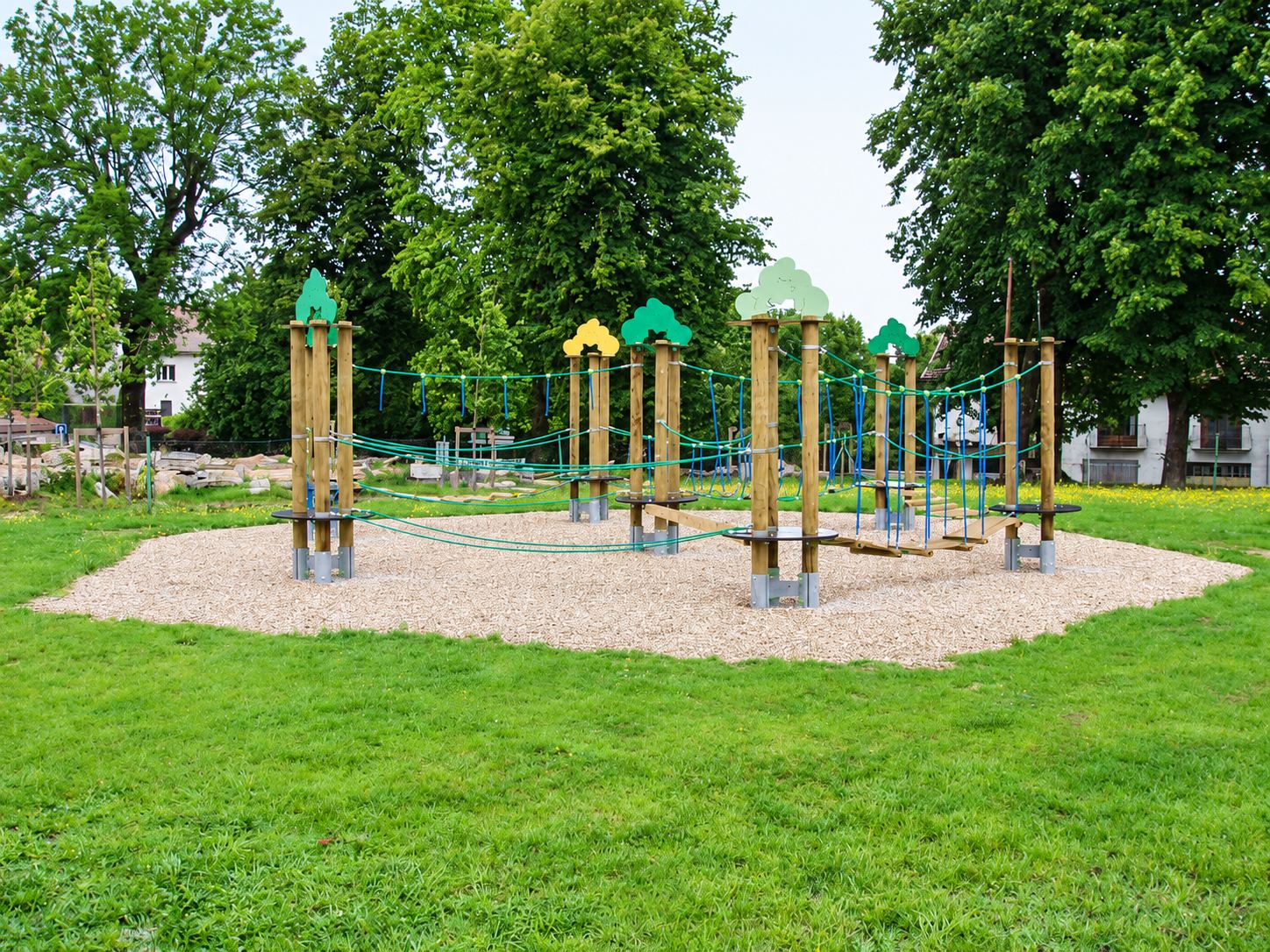Une structure de jeux en bois avec poteaux ornés de feuillages colorés, cordes et passerelles, installée sur un lit de gravier au milieu d'une pelouse entourée d'arbres.