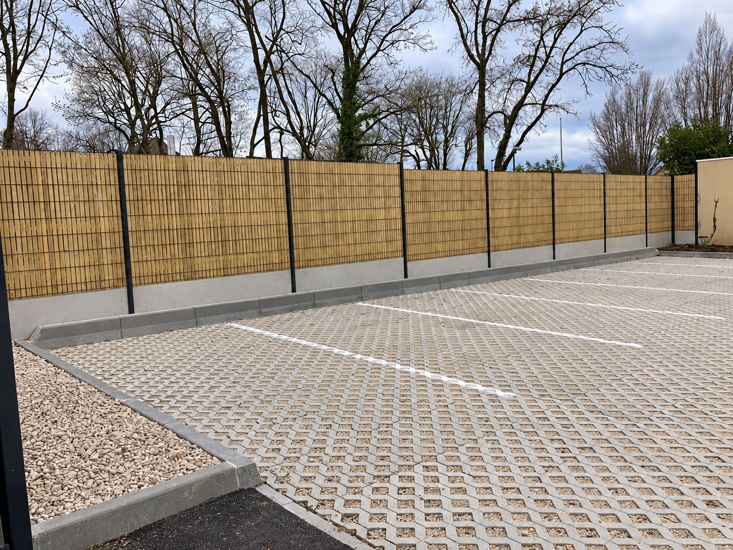Un parking récemment aménagé en dalles alvéolées bordé de graviers et d'une clôture en lames de bois sur un muret en béton.