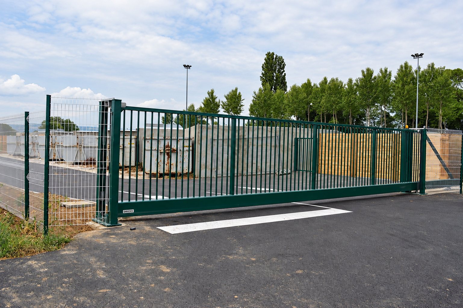 Un grand portail coulissant vert à barreaux verticaux, entouré de clôtures grillagées, ferme l'accès à un site avec des bennes et un parking en enrobé.