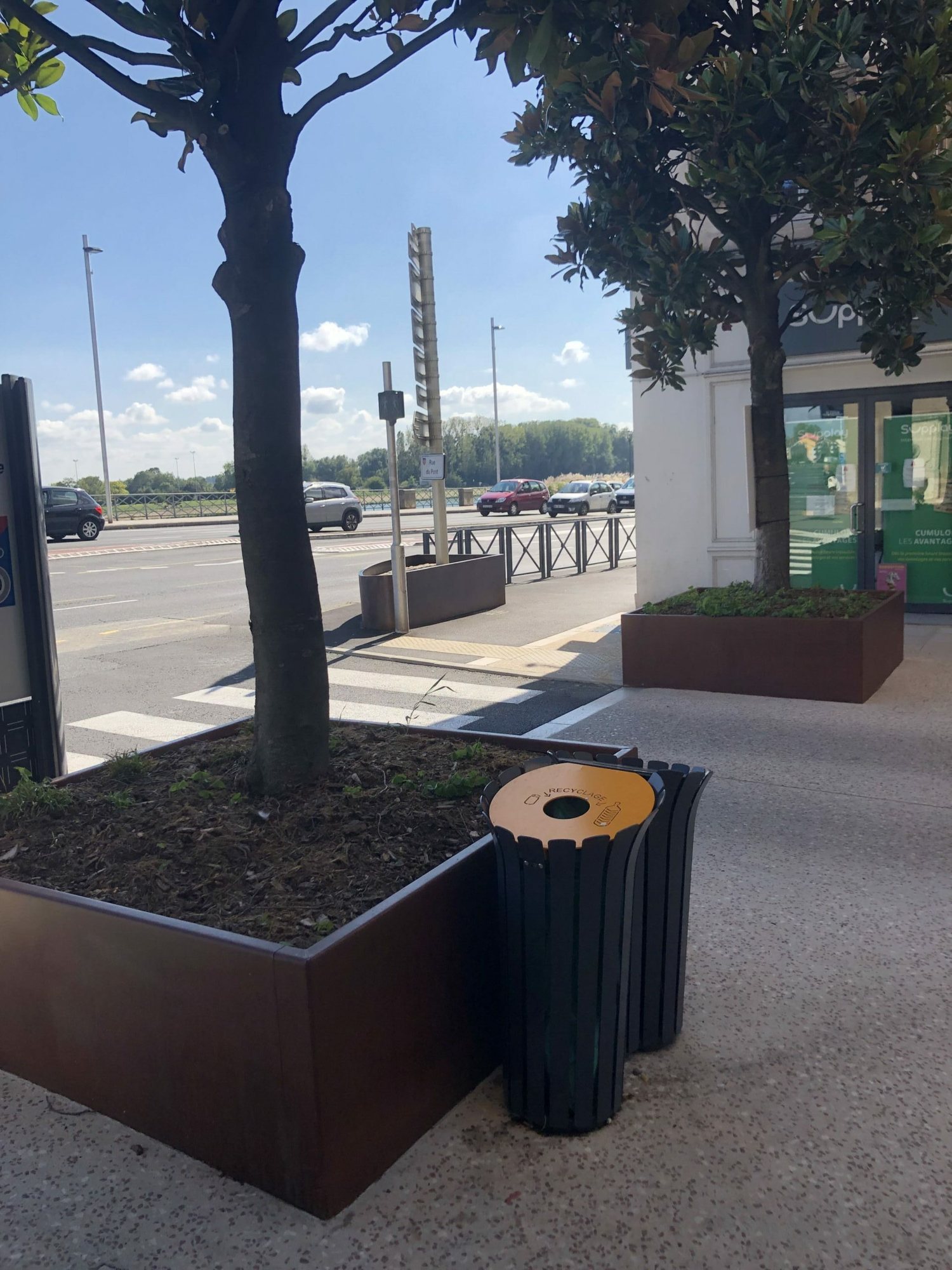 Une poubelle de recyclage noire à couvercle orange est posée sur un trottoir, à côté de grands bacs en acier corten plantés de magnolias, devant une rue et une boutique.