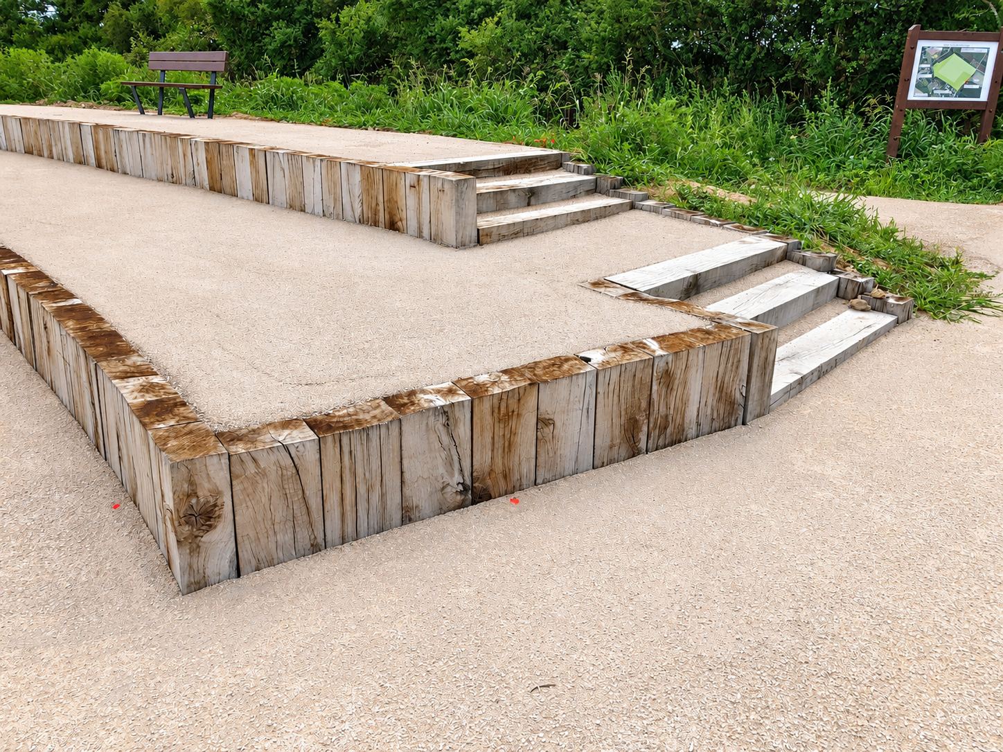 Une terrasse en gravier stabilisé entourée de traverses de bois verticales, avec deux petits escaliers en bois, un banc et un panneau d'information en arrière-plan.