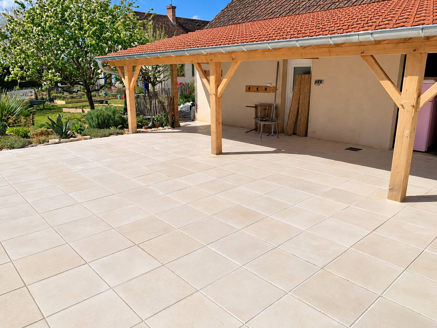 Une grande terrasse carrelée de dalles beige clair s'étend sous un auvent en bois couvert de tuiles rouges, devant un jardin fleuri.