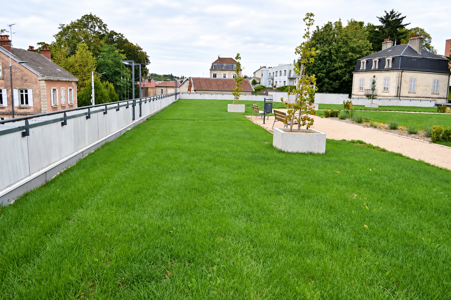 Une vaste pelouse verte aménagée sur une terrasse, bordée d'un garde-corps, avec des bancs, de jeunes arbres plantés dans des bacs en béton et des maisons en arrière-plan.